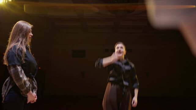 Rehearsal on dimly lit stage with performers practicing expressive gestures and interactions under warm theatrical lighting in an intimate empty theater setting