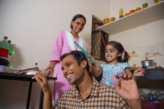 Little girl combing her father's hair in the kitchen while mother standing behind