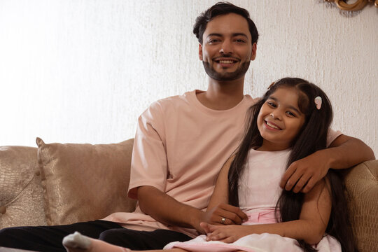 Father and daughter looking at camera while sitting together on sofa at home