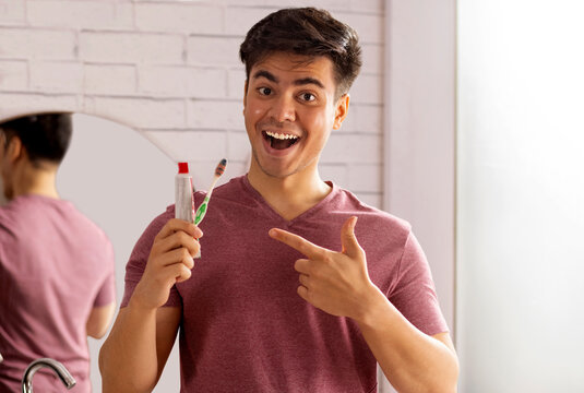 Cheerful young man pointing at toothpaste and toothbrush holding in his hand 
