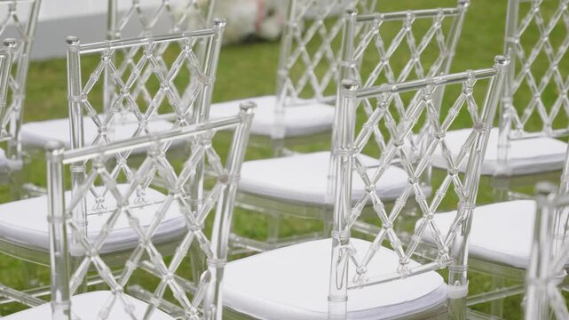 Clear acrylic chairs with white cushions are neatly arranged for a wedding ceremony in a beautiful outdoor garden setting