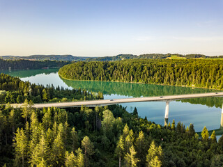 Obraz premium Aerial view of a long concrete viaduct bridge crossing the emerald green Lech river winding through dense forests in Bavaria, Germany.