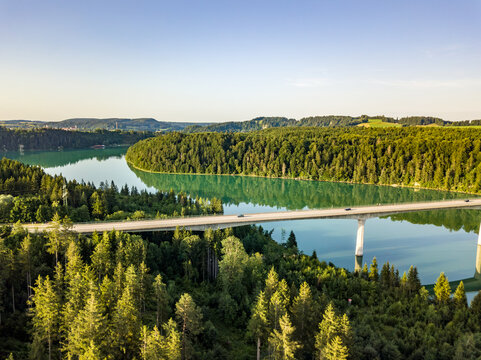 Aerial view of a long concrete viaduct bridge crossing the emerald green Lech river winding through dense forests in Bavaria, Germany.