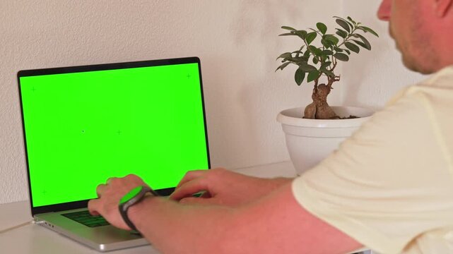 A person sits at a desk with a laptop and a plant. They focus on the screen and type on the keyboard. The setting is simple and bright. They are learning software skills at home.
