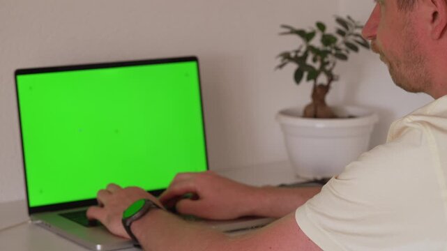 A person sits at a desk and types on a laptop that has a green screen displayed. There is a plant in the background. The setting is indoors, with a simple and clean look.
