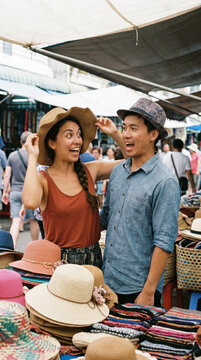 Laughing couple playfully trying colorful hats at a crowded outdoor artisan street market