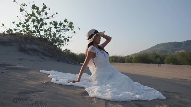 Young Indonesian southeast asian woman sitting on a sandy dune. She wears a flowing white dress and a straw sun hat, leaning back on one hand with the other touching her hat