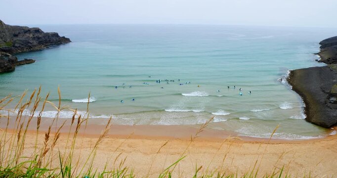 Surfer's Paradise: A breathtaking aerial view of a secluded cove showcases surfers catching waves under a soft, overcast sky, with rugged cliffs framing the golden sandy beach and turquoise waters.