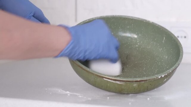 Blue gloved hand wipes darkened speckled frying pan with white sponge in bright kitchen sink area, showing careful household cleaning beside soap bottle and soft wall outlet background