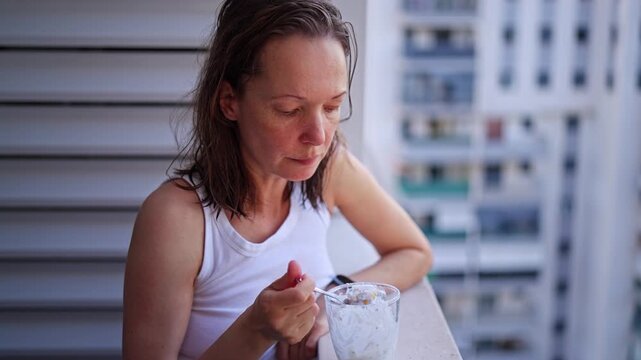 A woman sits on a balcony and enjoys ice cream on a warm summer day. She takes bites with a spoon while smiling and having a good time. The atmosphere is cheerful.