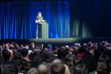 Large audience watching presenter on stage during technology conference. Blue curtains, spotlight, cameras, phones capturing keynote, focused crowd, professional atmosphere. © Anton Gvozdikov