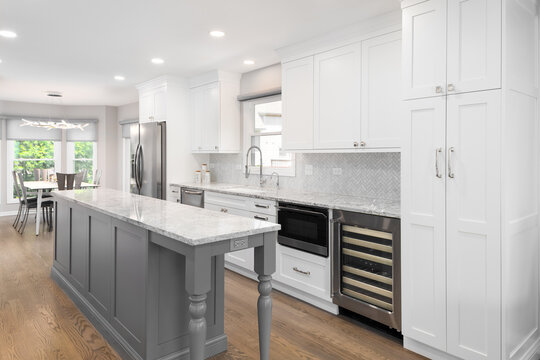 A modern kitchen with white cabinets, grey countertop, and a herringbone tile backsplash over a marble countertop looking towards a dining room.