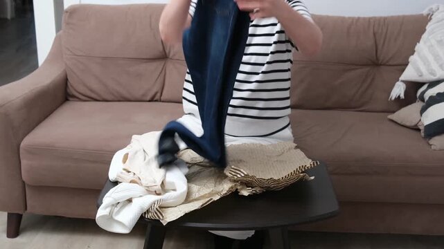 Woman open package and watching jeans outward sorting on sofa in cozy living room with small table, folded clothes, and calm daylight, with woman, sorting, folding detail.