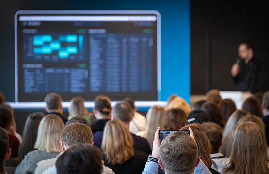 Conference audience watching presenter and projected data screen, attendee records presentation on smartphone, professional meeting atmosphere.