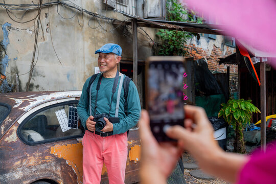 Older man poses for a picture in front of an old car in a rundown area during daylight