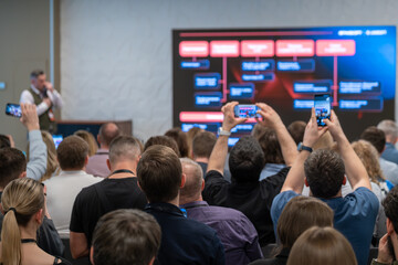 Obraz premium Conference audience records business presentation on large display, attendees capture slides with smartphones, focused atmosphere, learning and networking.