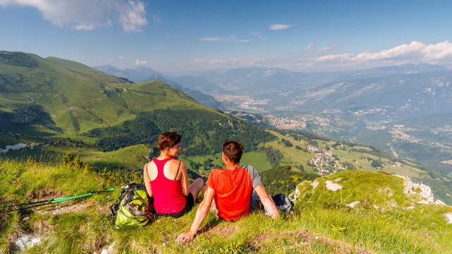 Active couple hiking sits on a mountain cliff enjoying an expansive alpine vista, representing travel and adventure at Monte Baldo Natural Park, Brentonico, Trentino, Veneto, Italy.