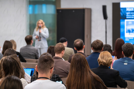 Business speaker presenting to attentive audience at indoor conference, professionals taking notes and using devices.
