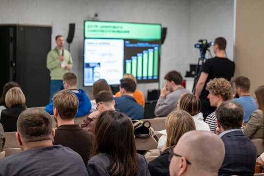 Business conference scene with presenter, seated audience, presentation screen and camera recording, professional networking and learning atmosphere.