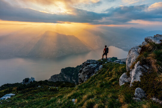 Hiker standing on mountain cliff watches sunset over lake, symbolizing adventure and travel freedom at Altissimo Refuge, Monte Baldo Natural Park, Brentonico, Trentino, Veneto, Italy.