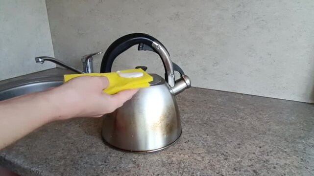 Hands scrubbing a silver stovetop kettle with a bright yellow sponge on a mottled grey countertop beside a stainless steel sink.