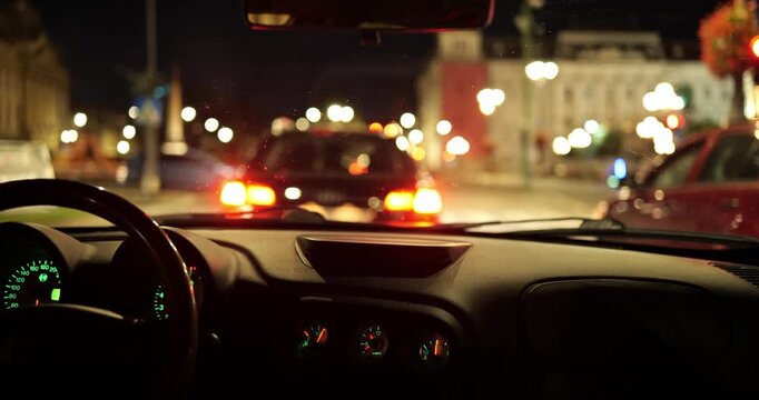 Car dashboard and steering wheel looking out at blurred city traffic lights while waiting at a red light, POV