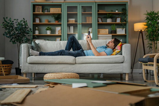 Young man procrastinating on sofa avoiding diy furniture assembly