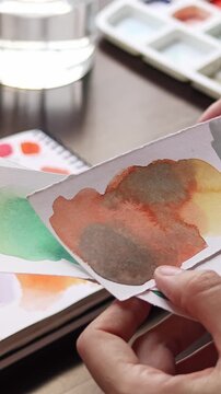 Artist examining watercolor color swatches on paper to compare paint mixtures before starting a painting. Creative process of selecting color combinations, testing pigments and planning. Close-up view