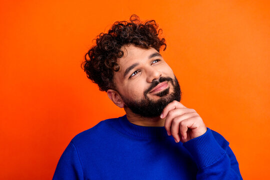 Thoughtful young man in blue sweater looking up with a smile on orange background, portrait of a bearded curly-haired person pondering an idea and feeling optimistic