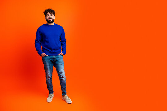 Smiling man in blue sweater standing against bright orange background, casual portrait with hands in pockets and relaxed confident expression