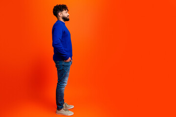 Young man standing in profile wearing blue sweater and jeans against a vibrant orange background, casual confident pose with hands in pockets and thoughtful expression © deagreez
