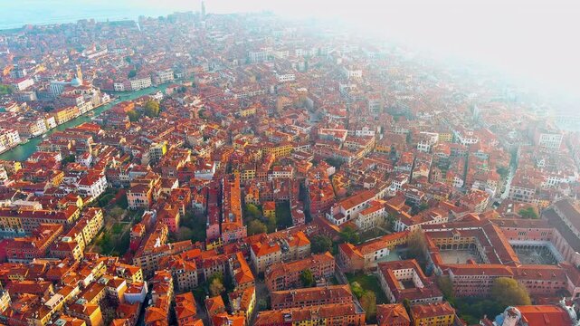 Aerial view of the Venice cityscape in Italy at sunset, with terracotta rooftops of the Cannaregio district, church domes, bell towers, the Grand Canal and the lagoon under haze.