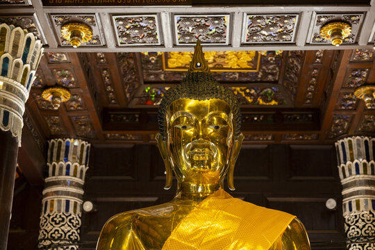 Golden Buddha statue in ornate temple interior with carved wooden ceiling and decorated columns, serene spiritual atmosphere and warm reflective light