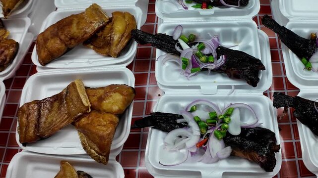Traditional grilled catfish served in takeaway food boxes with sliced red onion and fresh chili peppers at a Thai street food market. Authentic local cuisine prepared for customers and ready to eat. 4