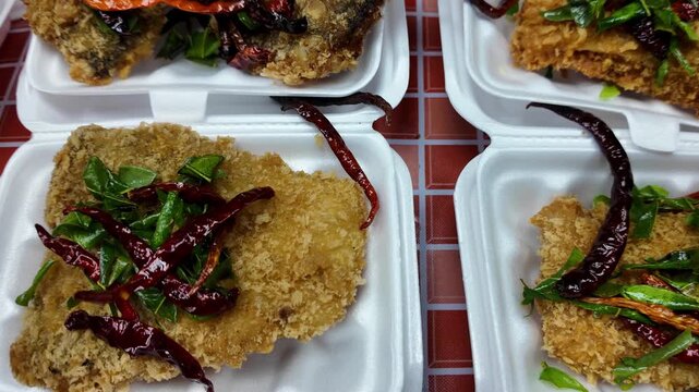 Close-up of crispy deep-fried fish served in takeaway food containers with fresh herbs and dried chili peppers at a Thai street food market. Traditional Thai seafood meal prepared and ready for custom