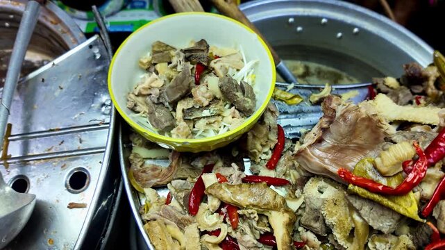 Close-up of traditional Thai pork offal including intestines, liver, skin and other cuts seasoned with red chili peppers and served from a large cooking pot at a busy street food market. Authentic loc