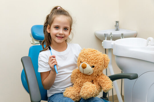 Little girl with long hair smiles while holding a teddy bear and a toothbrush, seated in dental chair within a well-lit dental clinic with sink and dental tools in the background. pediatric dentistry
