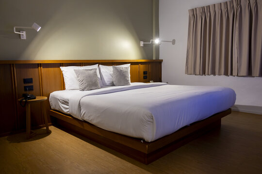 Modern minimalist hotel room with neatly made double bed, white linens, two decorative pillows, warm wooden headboard and soft lighting creating calm cozy atmosphere