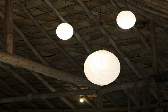 Paper lantern pendant light glowing in rustic wooden ceiling with exposed beams, warm ambient mood and textured thatch roof creating cozy architectural detail