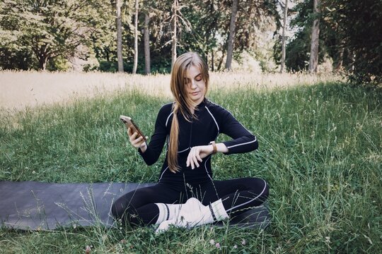 Young woman sits on yoga mat in grass looking at smartphone and smartwatch. Femtech, Technology for Womens Health, hormone health tracking, femtech wearable device.