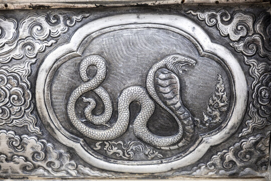 Silver bas relief panel with coiled cobra motif and decorative cloud border, intricate hammered metal texture conveying ancient temple craftsmanship and subtle dramatic mood