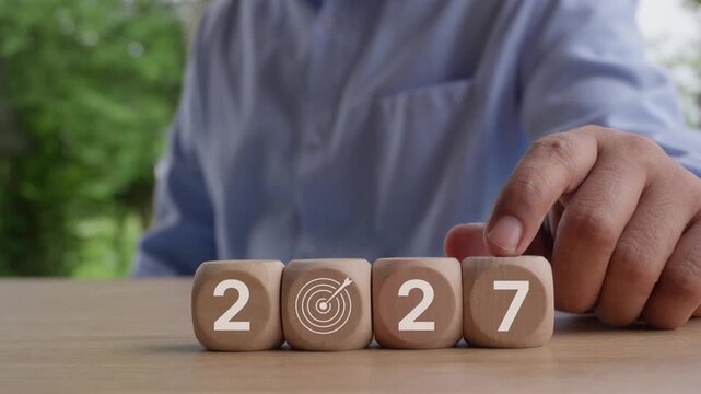 Business hand changing wooden cubes from 2026 to 2027 with target icon representing goal achievement, strategic planning, business growth, future success, annual objectives, and corporate vision