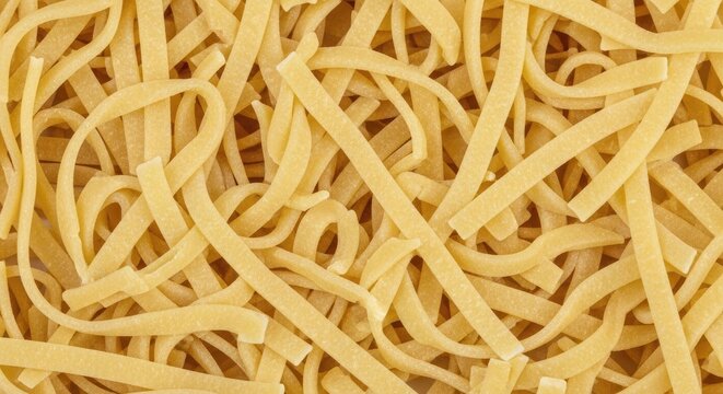A pile of fresh, uncooked fettuccine pasta lies on a dark surface. The pasta strands are a light golden color and are arranged in a tangled, haphazard manner.
