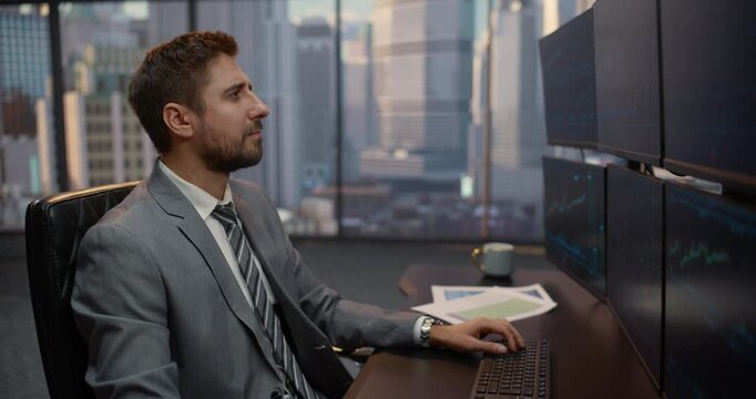 Portrait of Male Investor Studies Equity Indices on Multiple Monitors at His Desk, Analyzing Trading Data and Market Analytics to Forecast Capital Growth Opportunities. Big City Window View