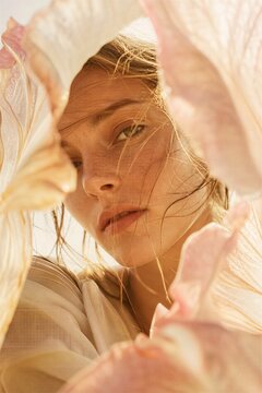 Young woman with freckles looking through delicate flower petals. Natural beauty and skincare concept. Portrait of girl behind translucent veil. Organic fashion and feminine aesthetic