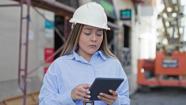 Woman architect in hardhat holding tablet and checking plans at building construction site; focused professionalism.