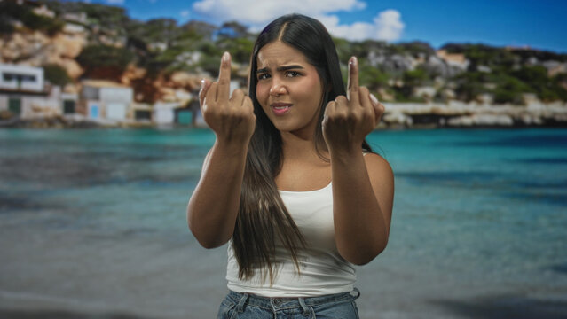 Woman showing middle fingers with both hands, grimacing while wearing white tank top and denim jeans at a sunny beach; defiance rebellion.