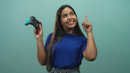Young woman gamer holding a teal and black gamepad, smiling and pointing index finger upward in studio with teal backdrop and blue shirt  playful confidence. © Krakenimages.com