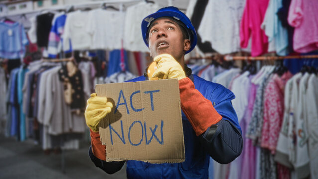 Man builder in hardhat holding cardboard sign reading act now amid clothing racks inside building wearing yellow gloves and blue uniform; urgent solidarity.