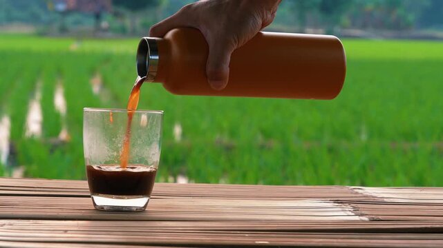 Hand pouring hot black coffee from orange thermal bottle into clear glass on wooden table with sunset field view